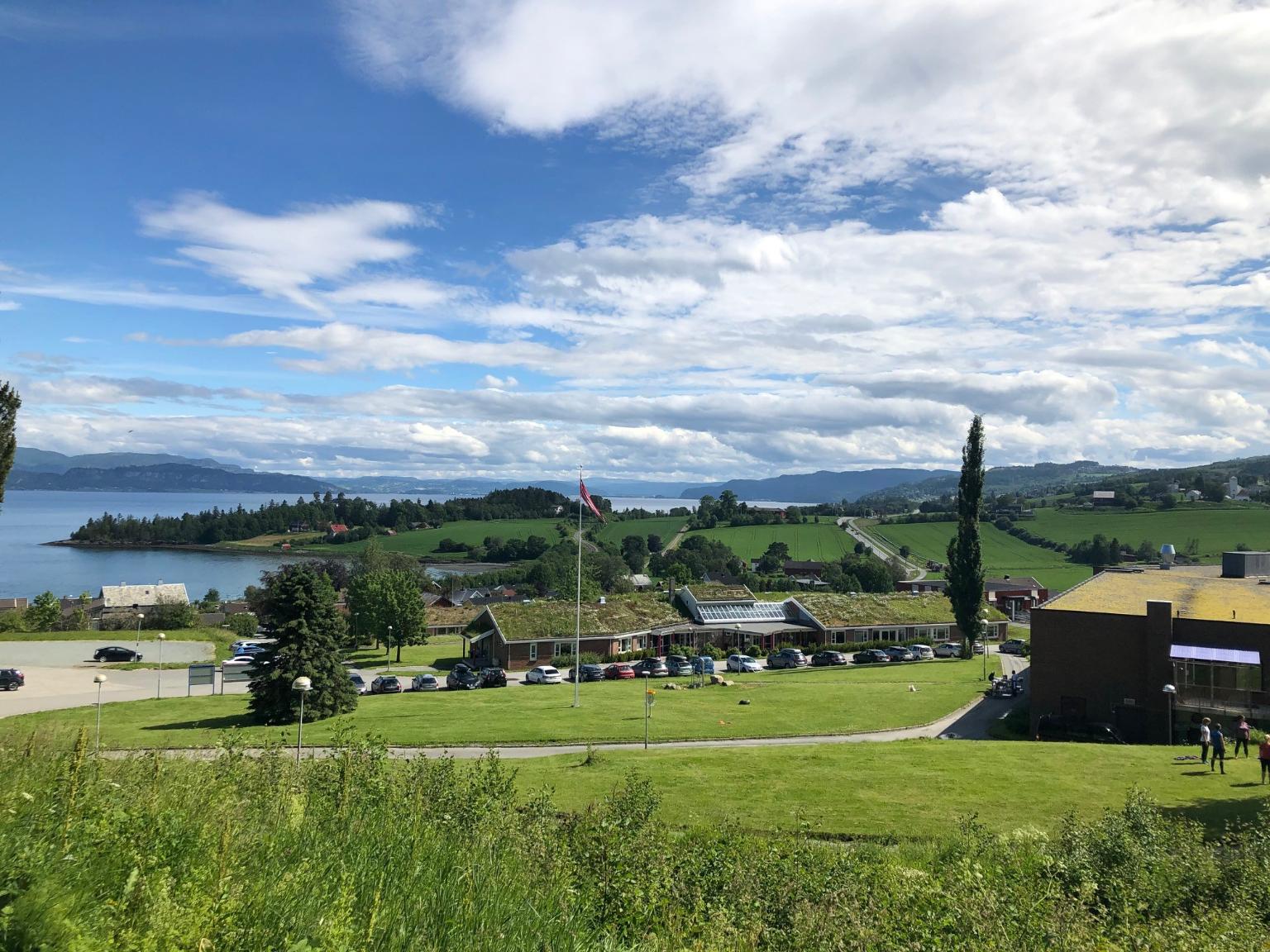 Utsikt over et grønt landskap med fjord, åkrer, bygninger med gress på taket og fjell i det fjerne.
